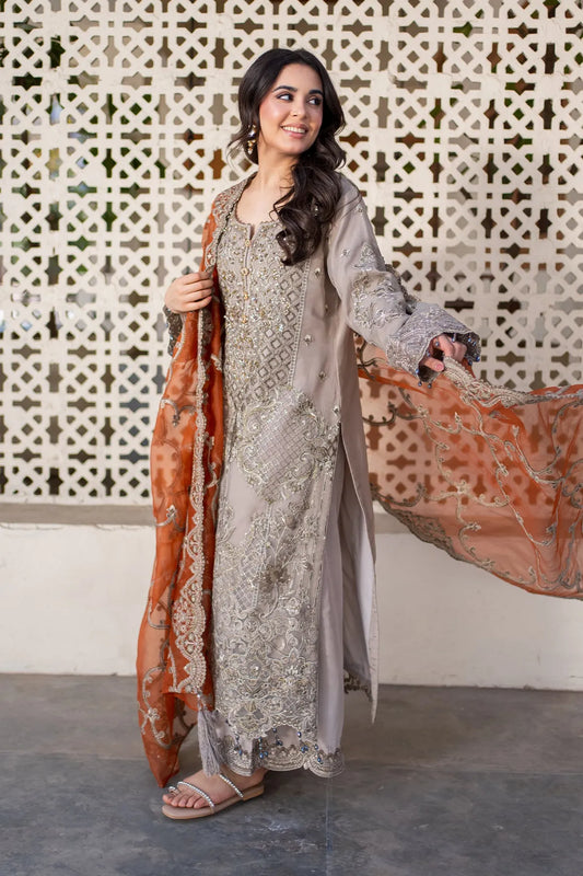 A woman wearing an elegant unstitched three-piece clothing ensemble with intricate embroidery, featuring a net dupatta in a rich, regal orange hue and a plain-dyed raw silk trouser, standing against a patterned backdrop.