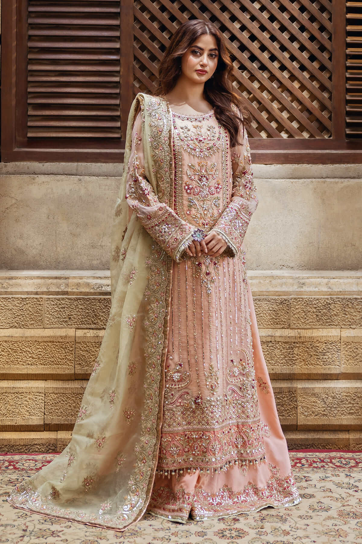 A woman wearing a traditional embroidered dress with peach and gold colors, featuring intricate handcrafted embroidery across the front, back, sleeves, and a dupatta.