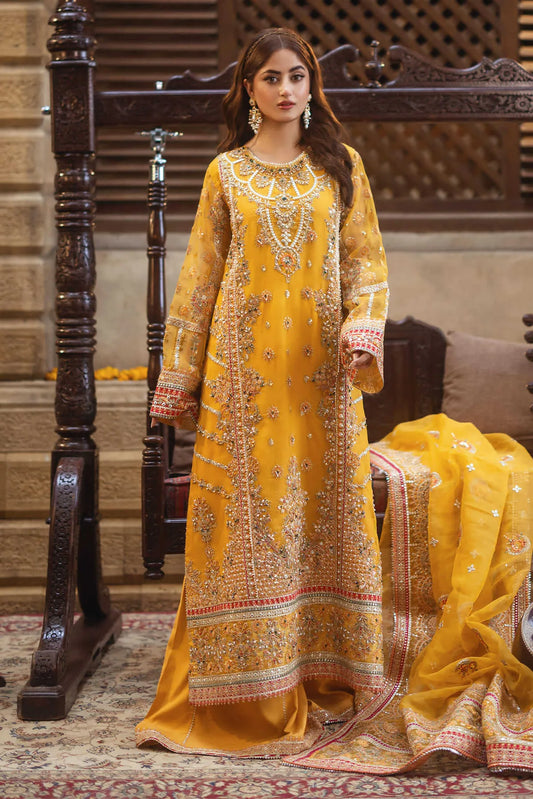 A woman wearing a traditional yellow embroidered gown with handmade embellishments, paired with a dupatta and sitting next to a couch with a decorative drape.