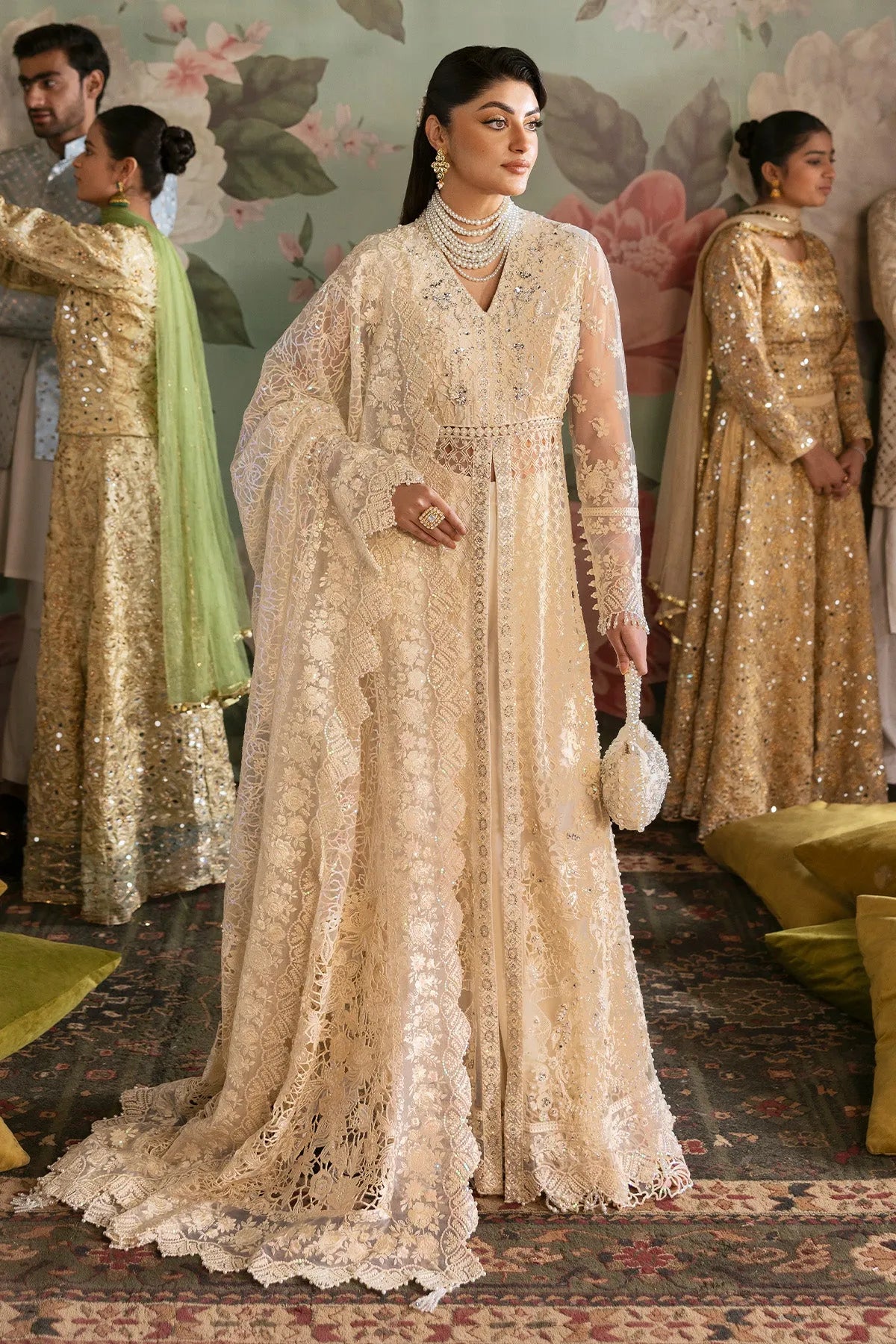 A woman wearing a traditional, embroidered beige gown with a lace bodice and net details, accompanied by a dupatta, in a room with other individuals dressed in similar attire.