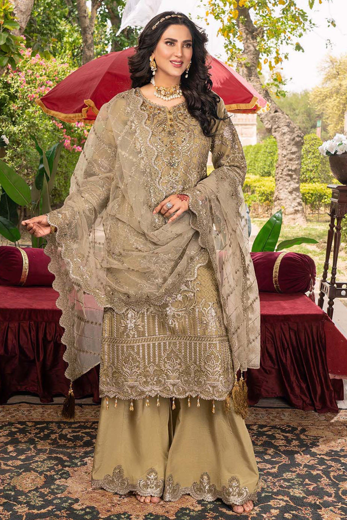 Woman in traditional embroidered outfit standing outdoors with a red umbrella and garden background