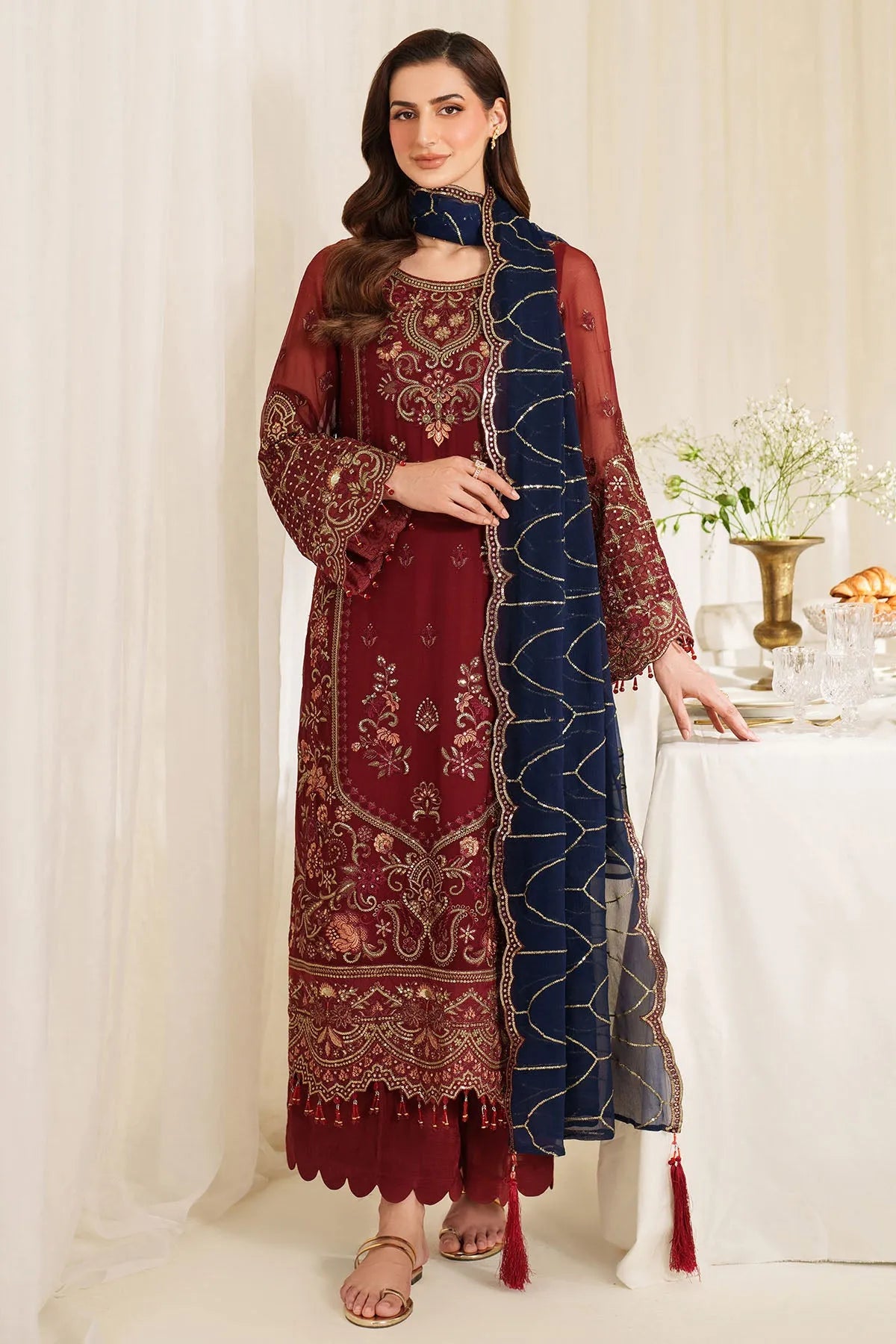Woman wearing a maroon and blue embroidered traditional outfit in a decorated room.